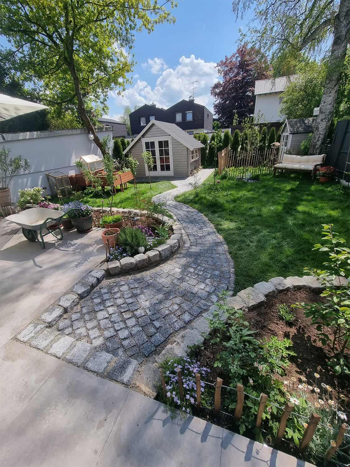 Ein liebevoll gestalteter Garten mit geschwungenem, gepflastertem Weg, der zu einem kleinen Gartenhaus führt. Der Garten ist mit verschiedenen Blumenbeeten, einem Hochbeet und einem Sitzbereich angelegt. Im Hintergrund stehen große, gepflegte Hecken, die den Garten umrahmen. Gartenbau und Landschaftsbau in München.