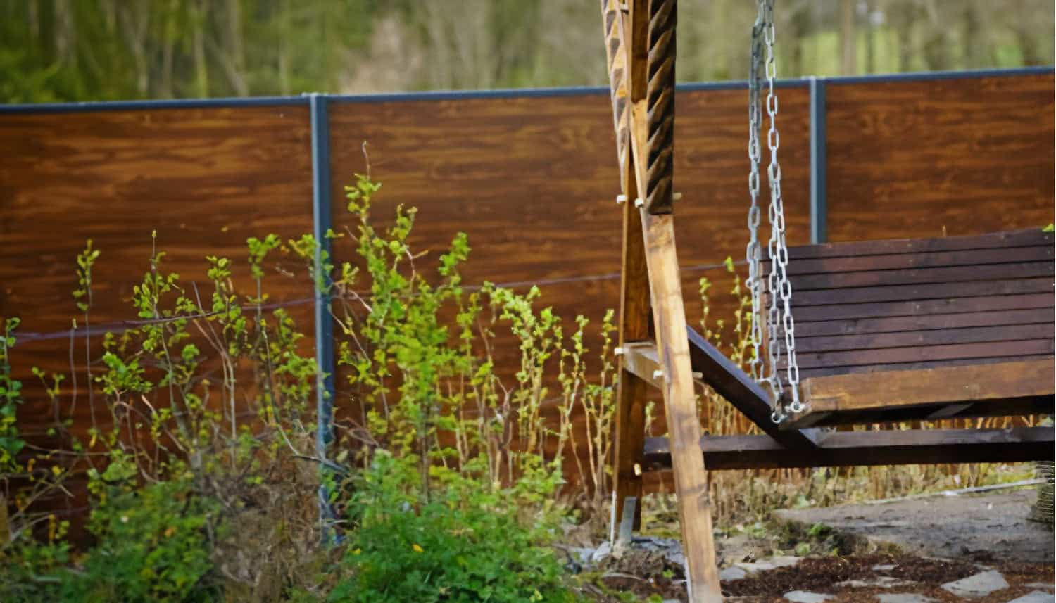 Ein Holzzaun dient als Sichtschutz im Garten. Im Vordergrund steht eine Holzschaukel, die an einer Metallkonstruktion befestigt ist. Dahinter wachsen einige Sträucher und Pflanzen.