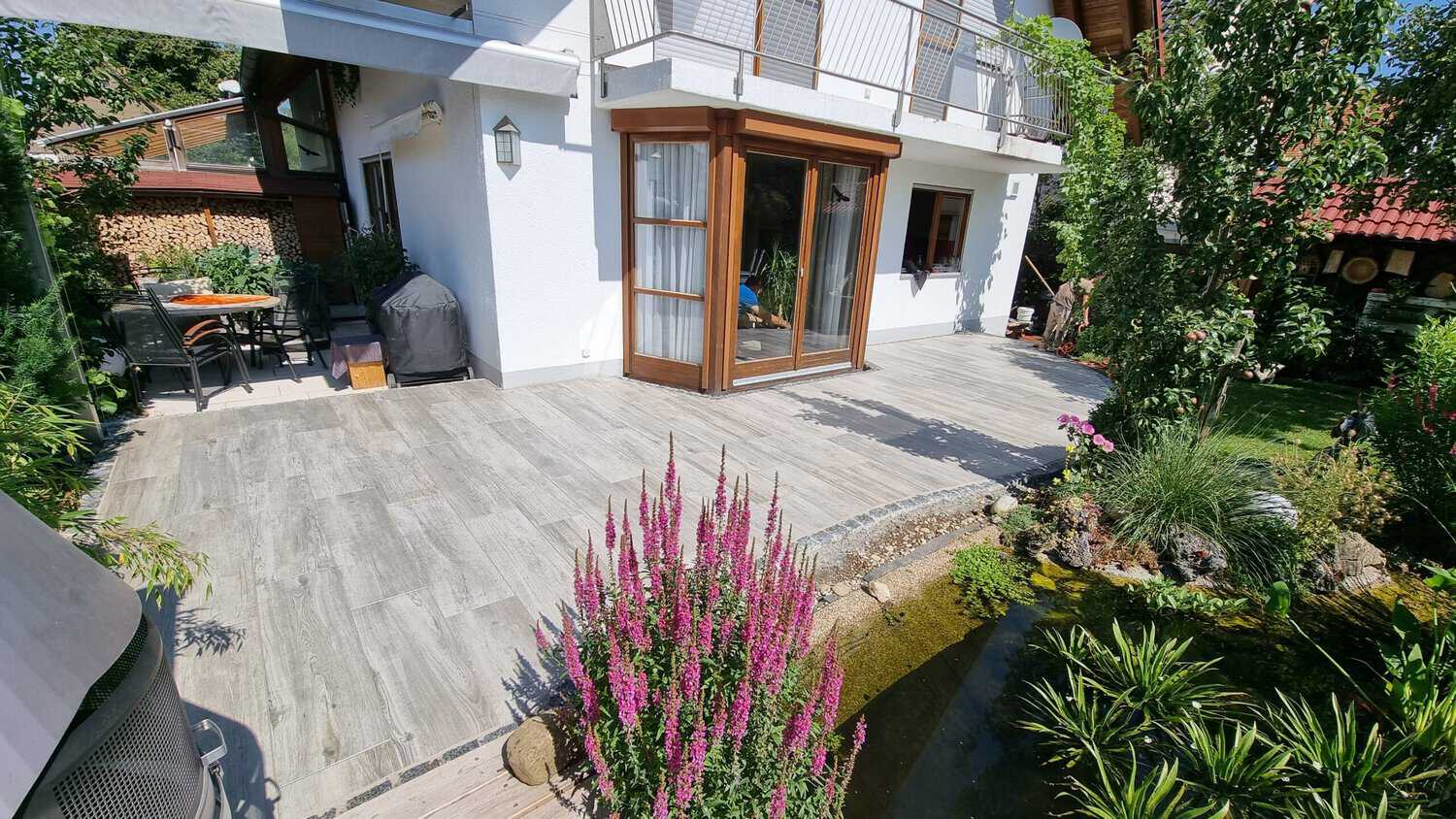 Terrassenbereich eines modernen Hauses mit Holzoptik-Fliesen, umgeben von einem gepflegten Garten mit einem kleinen Teich und blühenden Blumen. Im Hintergrund stehen Gartenmöbel sowie eine Grillstation, während das Haus mit großen Fenstern und einem Balkon gestaltet ist. Gartenbau & Landschaftsbei in München