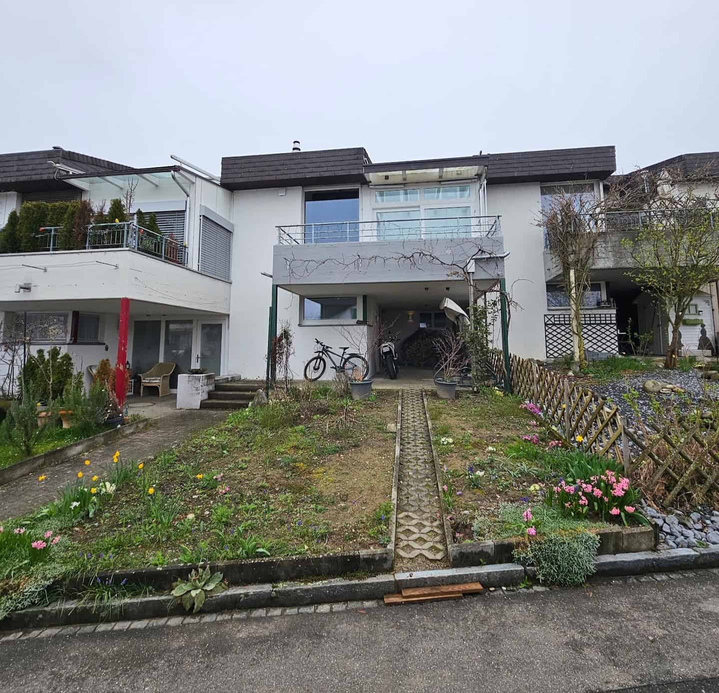 Ein liebevoll gestalteter Garten mit geschwungenem, gepflastertem Weg, der zu einem kleinen Gartenhaus führt. Der Garten ist mit verschiedenen Blumenbeeten, einem Hochbeet und einem Sitzbereich angelegt. Im Hintergrund stehen große, gepflegte Hecken, die den Garten umrahmen. Gartenbau und Landschaftsbau in München.