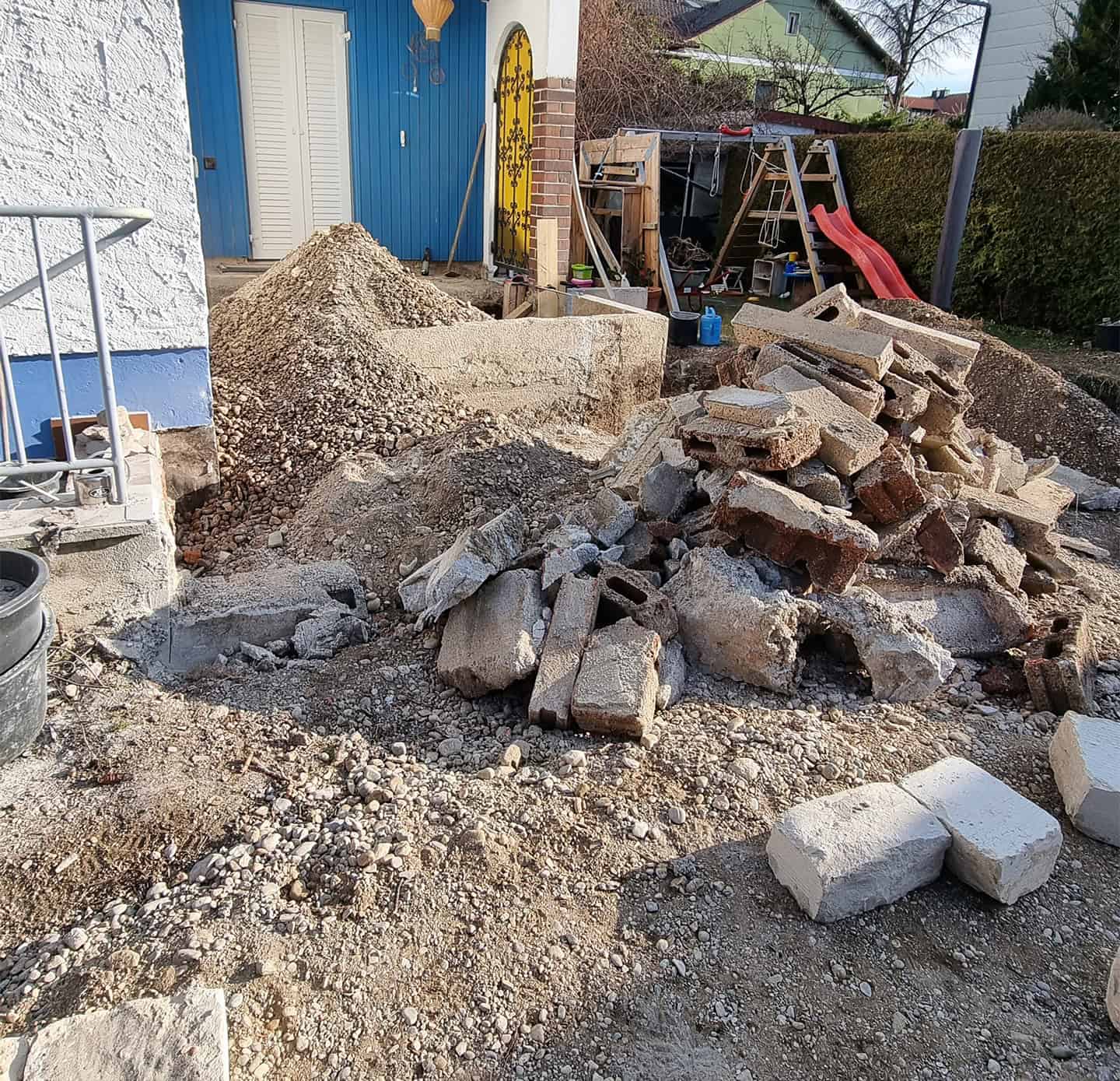 Ein liebevoll gestalteter Garten mit geschwungenem, gepflastertem Weg, der zu einem kleinen Gartenhaus führt. Der Garten ist mit verschiedenen Blumenbeeten, einem Hochbeet und einem Sitzbereich angelegt. Im Hintergrund stehen große, gepflegte Hecken, die den Garten umrahmen. Gartenbau und Landschaftsbau in München.